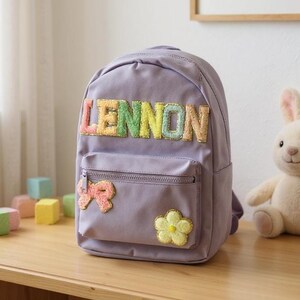 May include: A lavender backpack with the name "LENNON" in colorful, textured letters. The backpack has a front pocket with a bow and flower applique. A plush bunny toy is visible in the background.