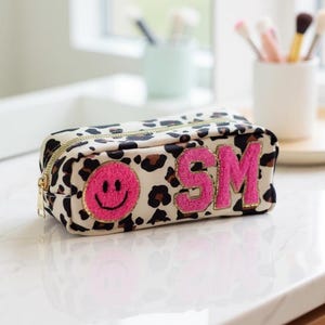 Könnte beinhalten: Eine rechteckige Kosmetiktasche mit cremefarbenem Leopardenmuster. Die Tasche hat einen goldenen Reißverschluss und ist mit einem rosa Smiley und den Buchstaben "SM" in Rosa mit goldfarbenem Glitzersaum verziert. Die Tasche liegt auf einer weißen Oberfläche.