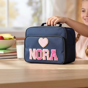 Op de afbeelding: Marineblauwe lunchtas met een roze hart en de naam "NORA" in roze letters. De tas heeft een zwart handvat en een voorvak. Een fruitschaal en een glas melk staan op de achtergrond.