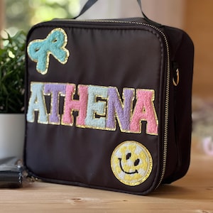 Könnte beinhalten: Eine schwarze Lunchtasche mit einem oberen Griff und einem goldenen Reißverschluss. Die Tasche trägt den Namen "ATHENA" in bunten, strukturierten Buchstaben, eine hellblaue Schleife und einen gelben Smiley-Aufnäher. Die Tasche hat eine goldene Umrandung.