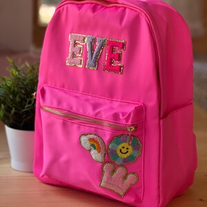 Può includere: Zaino rosa acceso con cerniere dorate e il nome "EVE" applicato sul davanti. Lo zaino ha una tasca frontale ed è decorato con toppe tra cui un arcobaleno, un fiore sorridente e una corona.
