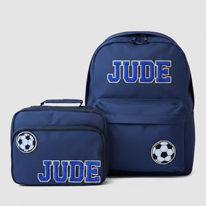 May include: A navy blue backpack and a matching lunch bag set. Both items feature the name JUDE in blue and white block letters and a black and white football patch. The backpack has a large main compartment and a front pocket with zippers.