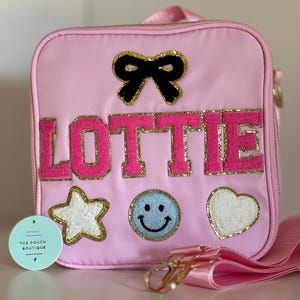 May include: Pink lunch bag with the name "LOTTIE" in pink and gold letters. The bag has a black bow, a white star, a blue smiley face, and a white heart. The bag has a pink strap and gold hardware.