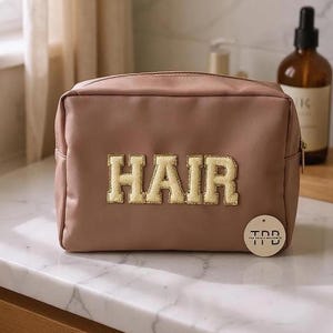Può includere: Un beauty case malva con la scritta "HAIR" in lettere ricamate dorate. La borsa ha una chiusura a zip e un piccolo tag rotondo con testo. Due bottiglie marroni sono sullo sfondo.
