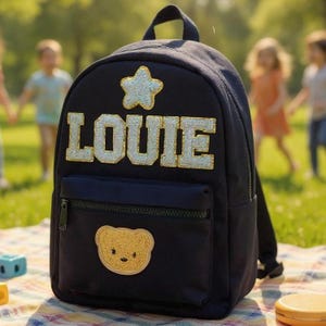 Puede incluir: Mochila azul marino con el nombre "LOUIE" en letras doradas con purpurina y una estrella dorada con purpurina. También se ve un pequeño bolsillo delantero y un parche de oso dorado. La mochila tiene un asa superior y correas ajustables.