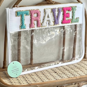 Könnte beinhalten: Weiße transparente Reißverschlusstasche mit goldenem Reißverschluss und dem Wort "TRAVEL" in bunten, flauschigen Buchstaben. Die Tasche liegt auf einer geflochtenen Weidenoberfläche.