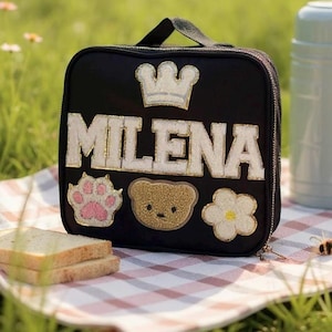May include: Black lunch bag with the name "MILENA" in white and gold lettering, a crown, paw print, teddy bear, and flower appliqués. The bag has a black handle and zipper. A water bottle and sandwich are in the background.