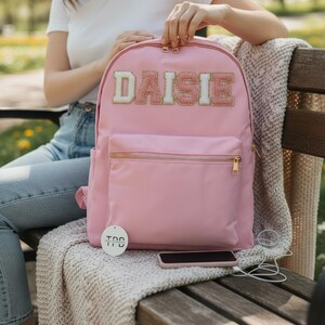 Può includere: Uno zaino rosa con una tasca frontale e cerniere dorate. Il nome "DAISIE" è ricamato sul davanti con un carattere decorativo. È attaccata una piccola etichetta bianca con testo nero. Uno smartphone e delle cuffie sono accanto allo zaino.