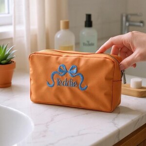 Op de afbeelding: Oranje rechthoekige cosmetische tas met een gouden rits en blauw geborduurde strik en de naam "Teddie". De tas staat op een wit aanrecht.