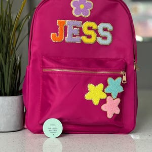 Può includere: Uno zaino rosa acceso con il nome "JESS" in lettere colorate e strutturate. Lo zaino presenta una tasca frontale con cerniera dorata e toppe a forma di stella. È visibile una piccola etichetta rotonda con testo.
