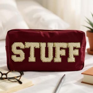 Può includere: Un astuccio rettangolare bordeaux con la scritta "STUFF" in lettere dorate e strutturate. L'astuccio è su una superficie bianca con un paio di occhiali, una penna e un libro nelle vicinanze. Una piccola pianta in vaso è sullo sfondo.