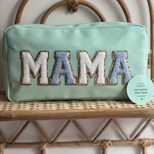 May include: A light green zippered pouch with the word "MAMA" in large, white, blue, and gold letters. The pouch is on a light brown wicker shelf.