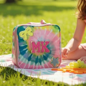 May include: A colorful tie-dye lunch bag with the initials "MK" in pink, gold, and blue. The bag has a handle and is decorated with gold and blue embellishments. The bag is sitting on a picnic blanket.