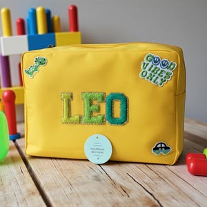 May include: A yellow pouch with the name "LEO" embroidered in green and blue. The pouch has a zipper closure and is decorated with patches including a dinosaur, a car, and the phrase "GOOD VIBES ONLY". The pouch is on a wooden surface.