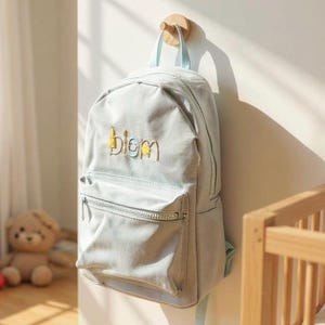 May include: A light blue backpack with a front pocket and a zipper closure. The word "blom" is embroidered in yellow and light blue on the front. The backpack is hanging on a wooden peg on a white wall.