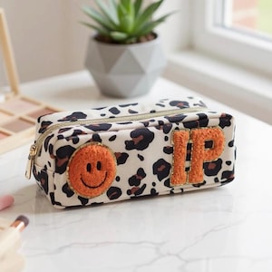 Könnte beinhalten: Eine rechteckige Kosmetiktasche mit einem Leopardenmuster in schwarzen und braunen Flecken auf cremefarbenem Hintergrund. Die Tasche hat einen goldenen Reißverschluss und einen orangefarbenen Smiley-Aufnäher. Die Buchstaben "IP" sind ebenfalls orange.