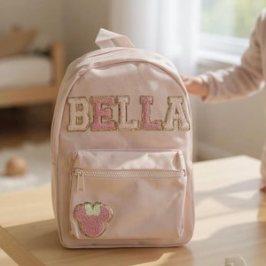 Può includere: Uno zaino rosa chiaro con il nome "BELLA" ricamato in lettere oro e rosa. Lo zaino ha una tasca frontale con cerniera e una piccola toppa di un orsetto rosa. Lo zaino è su una superficie di legno.