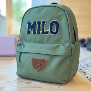 Peut inclure: Sac à dos vert olive avec le nom "MILO" en lettres universitaires bleu marine. Un écusson d'ours en peluche marron est cousu sur le devant. Le sac à dos a une poche avant et une poignée supérieure.