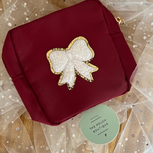 May include: A burgundy pouch with a white bow applique outlined in gold glitter. The pouch has a gold zipper and a tag that reads "THE POUCH BOUTIQUE". The background is a sheer, sparkly fabric.