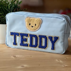 Könnte beinhalten: Eine hellblaue Kosmetiktasche mit dem Wort "TEDDY" in dunkelblauen, goldumrandeten Buchstaben. Ein kleiner, beigefarbener Teddybär-Aufnäher befindet sich über dem Text. Die Tasche hat einen Reißverschluss und ist wahrscheinlich aus Nylon.