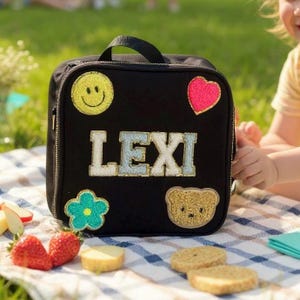 May include: Black lunch bag with a handle and zipper closure. The bag is decorated with a yellow smiley face, a pink heart, a blue flower, and a gold teddy bear. The name "LEXI" is embroidered on the front.