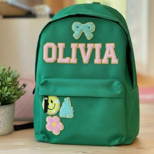 Può includere: Zaino verde con il nome "OLIVIA" in lettere rosa con bordo giallo. Lo zaino ha una tasca frontale ed è decorato con toppe tra cui un fiocco, una faccina sorridente, un fiore e una montagna.