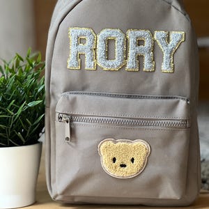 Può includere: Uno zaino grigio con il nome "RORY" in lettere bianche e dorate. Lo zaino ha una tasca frontale con cerniera e una toppa a forma di orsetto giallo.