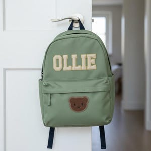 Può includere: Zaino verde con il nome "OLLIE" in lettere beige. Presenta una toppa a forma di orsetto marrone e spallacci neri. Lo zaino è appeso a una maniglia.