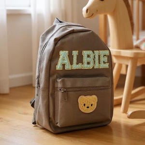 Può includere: Uno zaino marrone con il nome "ALBIE" in lettere verde chiaro e oro. Lo zaino ha una piccola tasca frontale con cerniera e una toppa a forma di orsetto. Un cavallo a dondolo di legno è sullo sfondo.