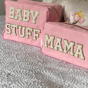Op de afbeelding: Twee roze canvas ritszakjes met gouden ritsen. Het grotere zakje heeft de tekst "BABY STUFF" in witte letters met gouden versiering. Het kleinere zakje heeft de tekst "MAMA" in witte letters met gouden versiering.