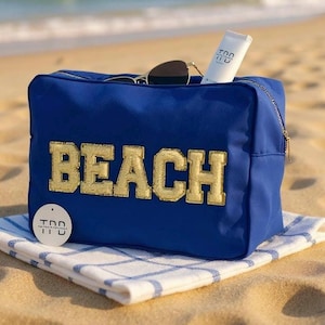 Pode incluir: Uma bolsa de praia azul com letras douradas "BEACH", óculos de sol e protetor solar. A bolsa está sobre uma toalha xadrez branca e azul em uma praia arenosa.