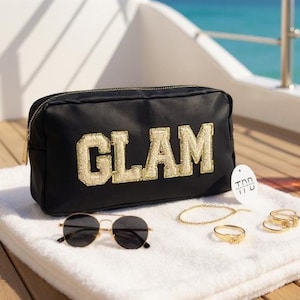 Può includere: Un beauty case nero con la scritta "GLAM" in oro, insieme a occhiali da sole con montatura dorata, un braccialetto d'oro e anelli d'oro. Gli oggetti sono disposti su un asciugamano bianco, suggerendo un tema di bellezza o viaggio.
