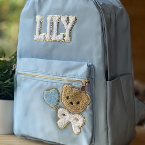 Könnte beinhalten: Hellblauer Rucksack mit dem Namen "LILY" in gold-weißen Buchstaben. Der Rucksack hat einen goldenen Reißverschluss und dekorative Aufnäher, darunter ein Herz, ein Teddybär und eine Schleife. Hergestellt aus einem glatten, strapazierfähigen Material.
