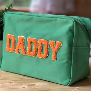 May include: A green rectangular cosmetic bag with the word "DADDY" in orange, glitter-edged letters. The bag has a zipper closure and is sitting on a wooden surface.