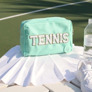 Può includere: Un beauty case verde menta con la scritta "TENNIS" ricamata in bianco e oro. La borsa è appoggiata su una gonna plissettata bianca. Nell'immagine sono presenti anche una bottiglia d'acqua e scarpe da ginnastica bianche.