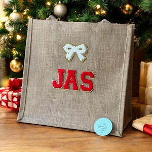 Può includere: Una borsa tote in iuta beige con le lettere "JAS" in rosso e un fiocco bianco con bordi dorati. La borsa è decorata per Natale e poggia su una superficie di legno.