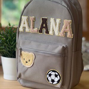Può includere: Zaino grigio con il nome "ALANA" in lettere beige, marroni e dorate. Presenta una toppa a forma di orsetto e una toppa a forma di pallone da calcio. Include una tasca frontale con cerniera.