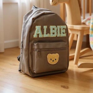 Può includere: Uno zaino marrone con il nome "ALBIE" in lettere verde chiaro. Lo zaino ha una tasca frontale con cerniera e una toppa a forma di orsetto giallo. È appoggiato su un pavimento in legno.
