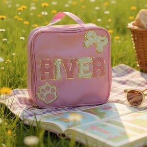 May include: Pink lunch bag with the name "RIVER" in gold and white letters. The bag has a bow and paw print applique. A handle is at the top. The bag is sitting on a picnic blanket with a book and sunglasses.