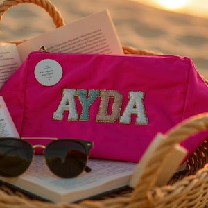 May include: A bright pink cosmetic bag with the name "AYDA" embroidered on it. The bag is inside a woven basket with a book and sunglasses. The bag has a zip closure and a small circular tag that reads "THE POUCH BOUTIQUE".