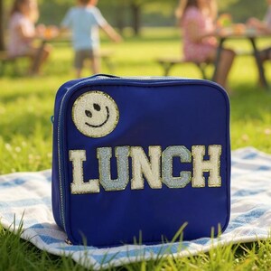 May include: A navy blue lunch bag with a smiley face patch and the word "LUNCH" in large, embroidered letters. The bag is sitting on a blue and white striped blanket on a grassy lawn.