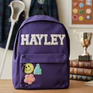 Könnte beinhalten: Ein lila Rucksack mit dem Namen "HAYLEY" in weißen Blockbuchstaben. Der Rucksack hat eine Vordertasche mit drei gestickten Aufnähern: einem lächelnden Gesicht, einem Berg und einer Blume. Ein Lacrosseschläger und eine Trophäe sind im Hintergrund.