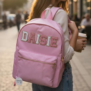 May include: A light pink backpack with the name "DAISIE" embroidered on the front. The backpack has a front pocket with a gold zipper and a top handle. The straps and the top handle are also light pink.