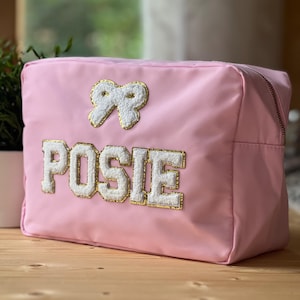 Op de afbeelding: Een roze rechthoekige make-up tas met een witte en gouden strik en de naam "POSIE" geborduurd op de voorkant. De tas heeft een ritssluiting en staat op een houten oppervlak.