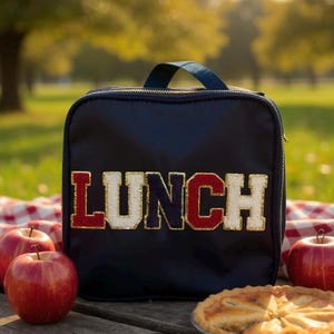 May include: A navy blue lunch bag with the word "LUNCH" in red, white, and blue varsity-style lettering. The bag has a handle and a gold zipper. Red apples and a pie are in the foreground.