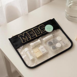 May include: A clear travel pouch with a black trim and the word "MEDS" in gold lettering. The pouch contains small white cylindrical containers, small square containers, and round containers. A small, round, green label is also visible.