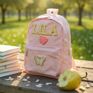 Puede incluir: Una mochila rosa con el nombre "LILA" en letras doradas con purpurina. La mochila tiene un corazón rosa y un diseño de mariposa. Libros y una pera sobre una superficie de madera.