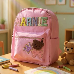 May include: A pink backpack with the name "MARNIE" embroidered in colourful letters. The backpack has a front pocket with a gold zip and patches of a bear, crown, and bow. A teddy bear is nearby.