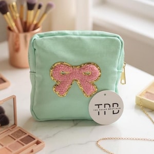 May include: A mint green cosmetic bag with a pink bow applique outlined in gold glitter. The bag has a gold zip and a white tag that reads "The Pough Boutique". Makeup brushes and eyeshadow palettes are in the background.