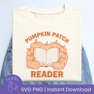 Könnte beinhalten: Cremefarbenes T-Shirt mit einem herbstlichen Design. Das Design zeigt einen Kreis aus Kürbissen, die ein offenes Buch umgeben, mit den Worten "PUMPKIN PATCH READER" in orangefarbenen Buchstaben. Das T-Shirt liegt auf einer Jeansoberfläche.